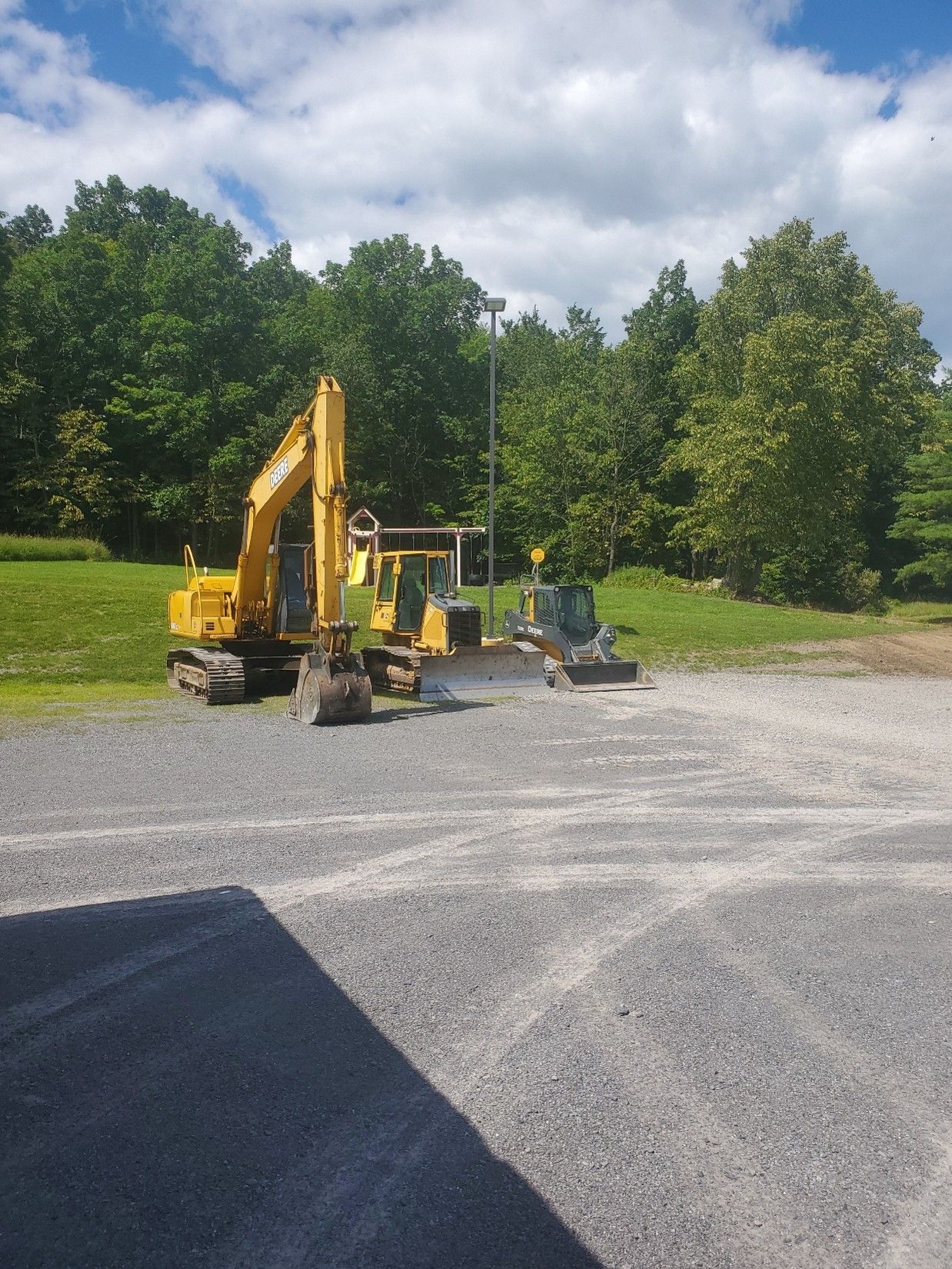 Yellow excavator and compact machine on a gravel lot with trees and a partly cloudy sky.
