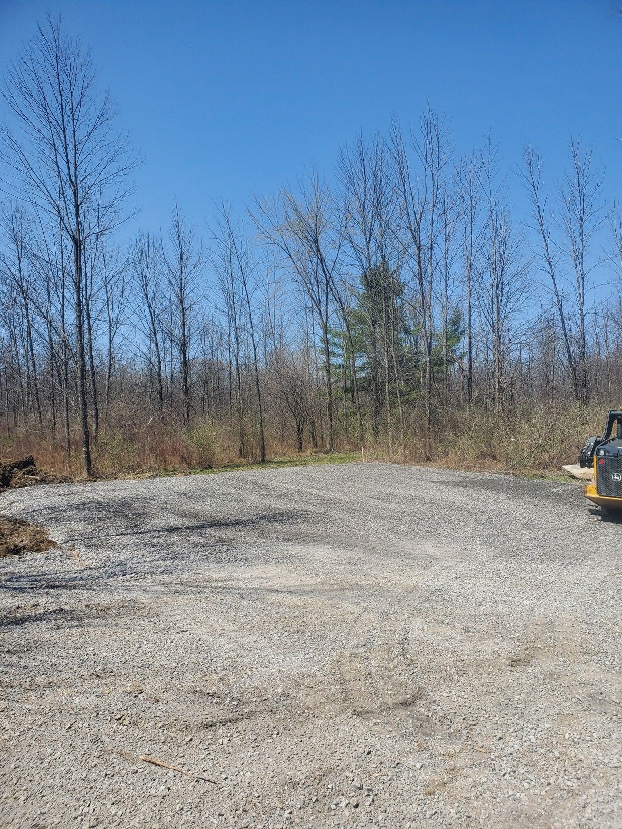 Gravel clearing beside leafless trees under a clear blue sky, with construction equipment at right.