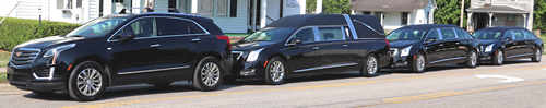 A procession of black hearses and vehicles parked on a street near a white building with green trees.