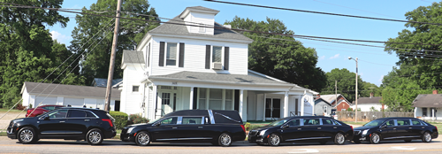 Funeral home with several black cars parked out front, including a hearse. White building, blue sky, trees.