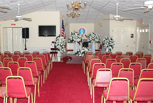 A funeral home chapel with rows of red chairs, altar with flowers, and an American flag.