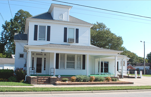 White two-story house with porch. Black shutters and a dormer. Brick and green shrubbery in the front yard.