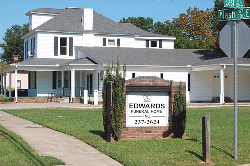 Edwards Funeral Home, Inc. building with sign on a grassy lawn; at street corner; white facade with black shutters.