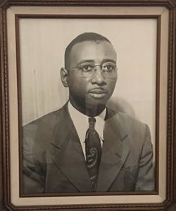 Black and white portrait of a man wearing glasses, suit, and tie in a framed photo.