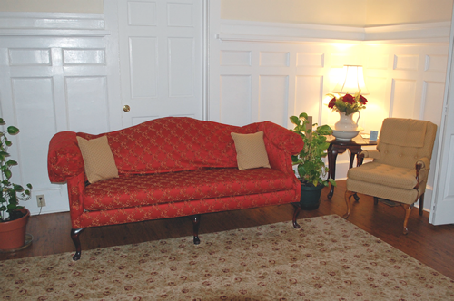 Red couch, tan chair, and rug in a room with white paneling and a table lamp.
