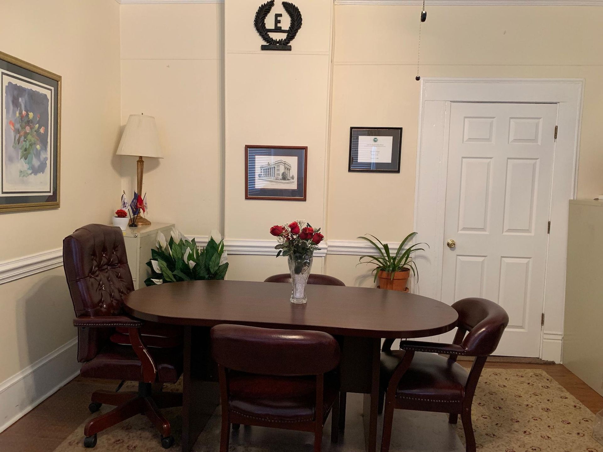 A conference room with a table, chairs, artwork, plants, and a door.