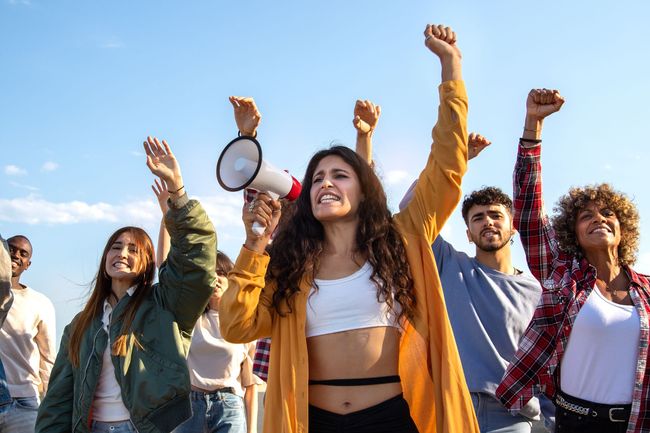 Group of people cheering outdoors with raised fists; one person holds a megaphone under a blue sky