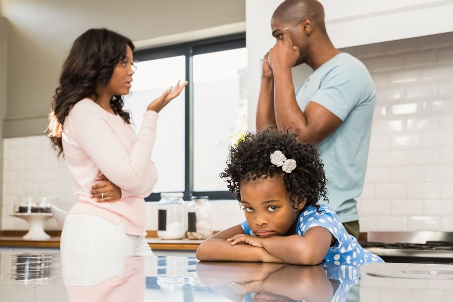 Three people in a kitchen, one child leaning on the counter while two adults talk nearby.
