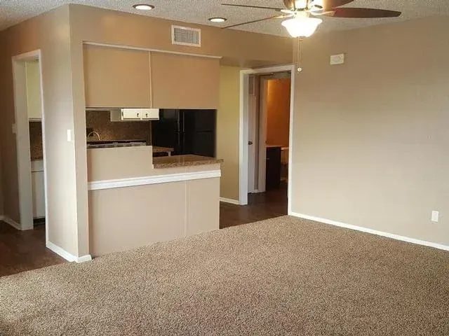 Empty living room with beige carpet, ceiling fan, and small kitchen pass-through.