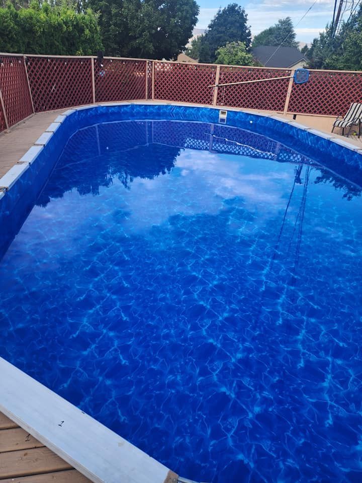 Blue pool in a backyard with a wooden deck and a lattice fence. Reflects sky.