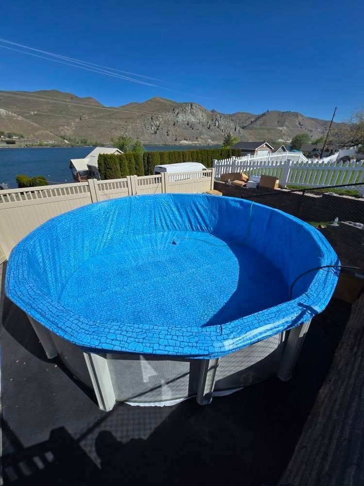 Above-ground pool covered with a blue tarp, overlooking a lake and mountains on a sunny day.