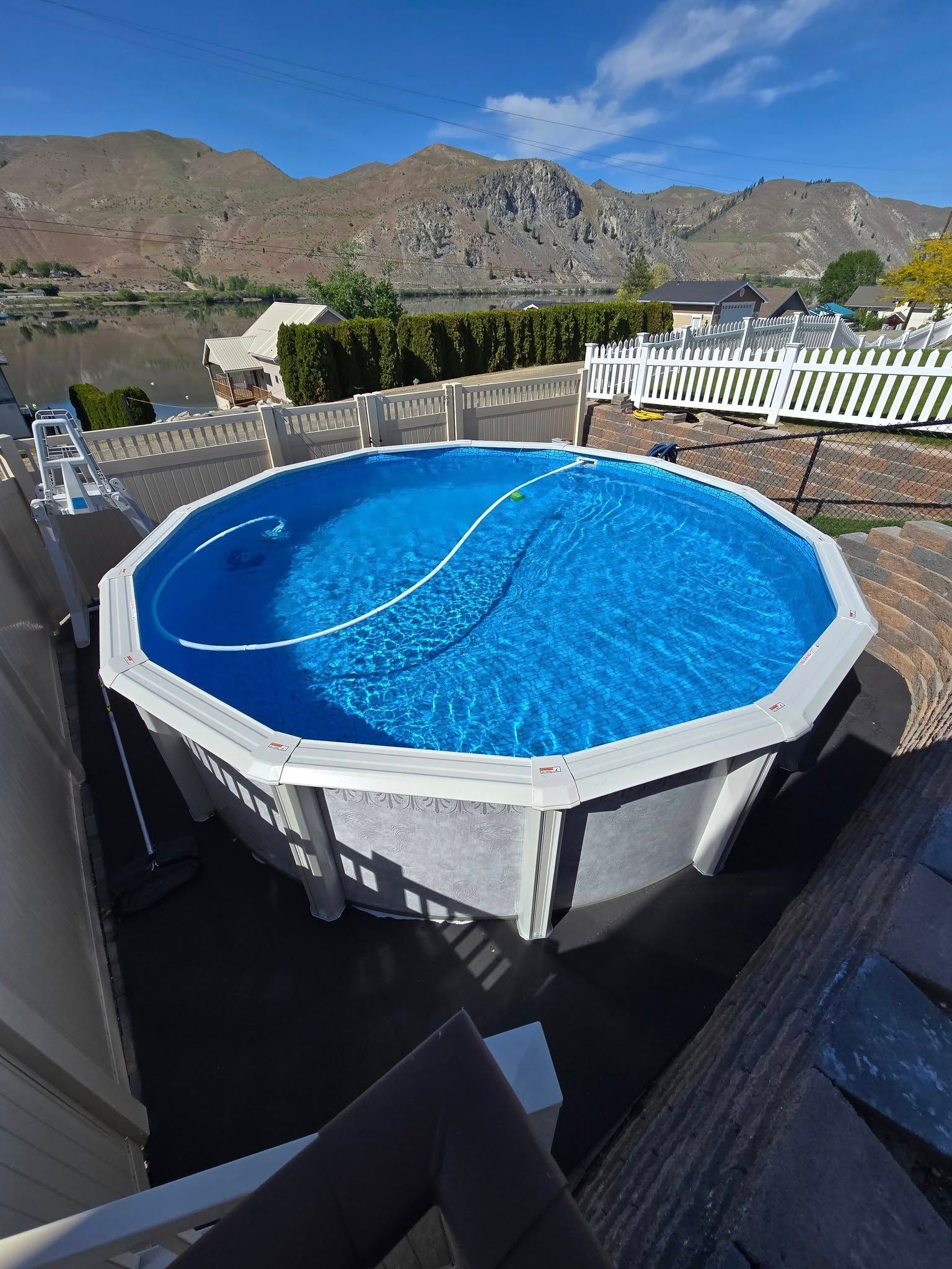 Above-ground pool filled with blue water, surrounded by a fence, with mountains in the background on a sunny day.