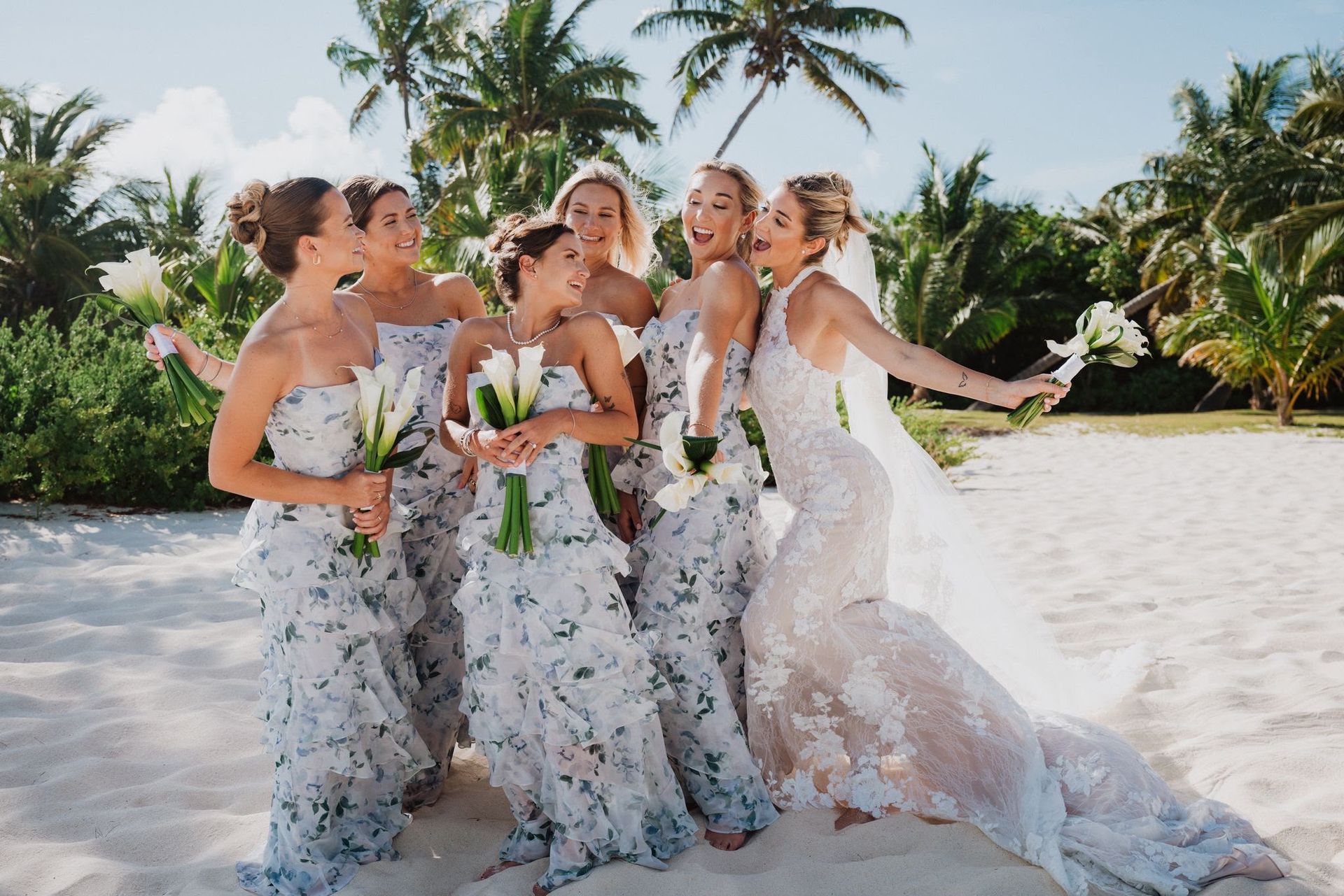 Five women in white floral dresses pose on a sunny beach with palm trees