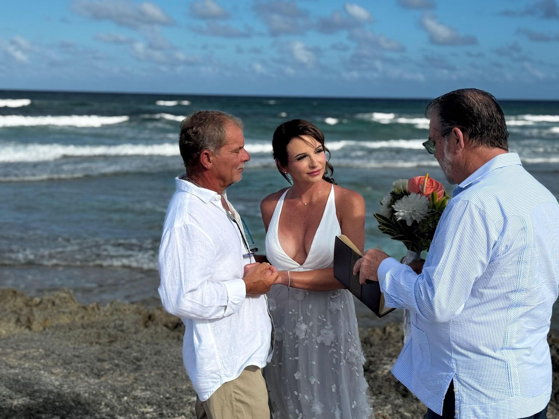 Three people stand on a beach during a small wedding ceremony, with waves in the background.