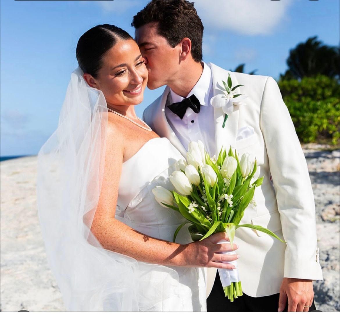 Bride and groom in white wedding attire embracing on a sunny beach, holding a bouquet