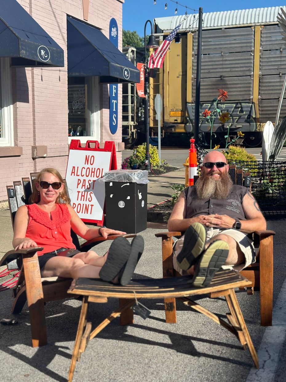 A man and a woman are sitting in wooden chairs on the sidewalk.