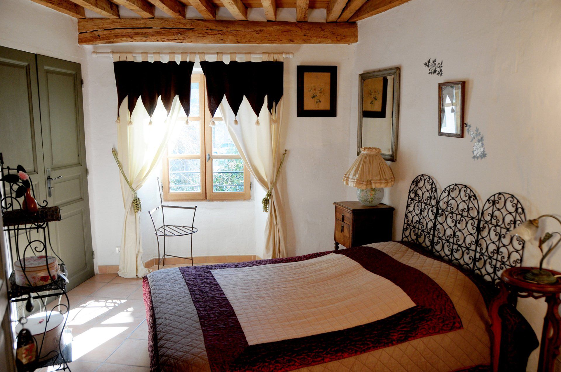 Chambre confortable avec un lit, une fenêtre à rideaux, une tête de lit à motifs et des poutres apparentes au plafond.