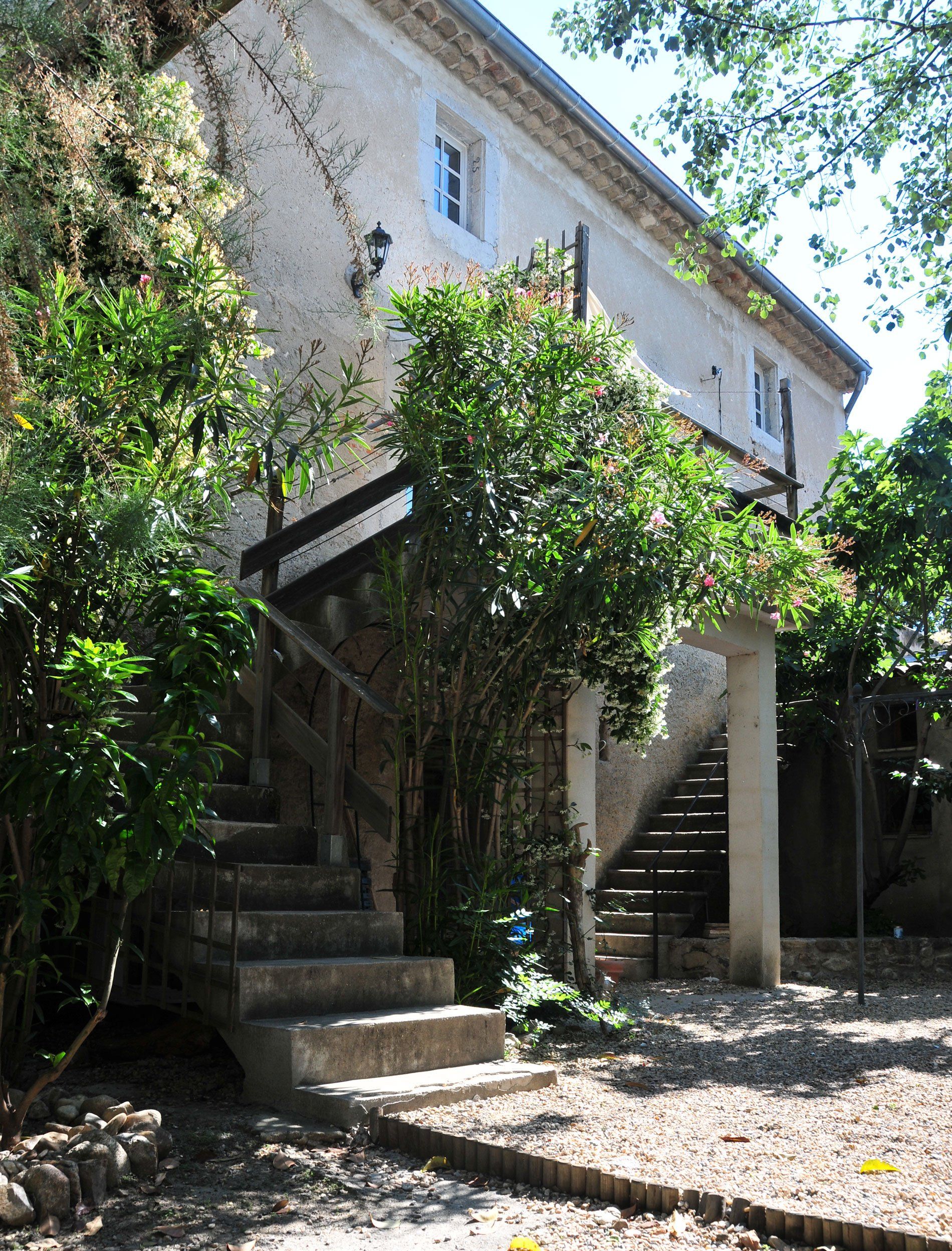 Maison en pierre avec escalier extérieur, entourée d'arbres verdoyants et de soleil.