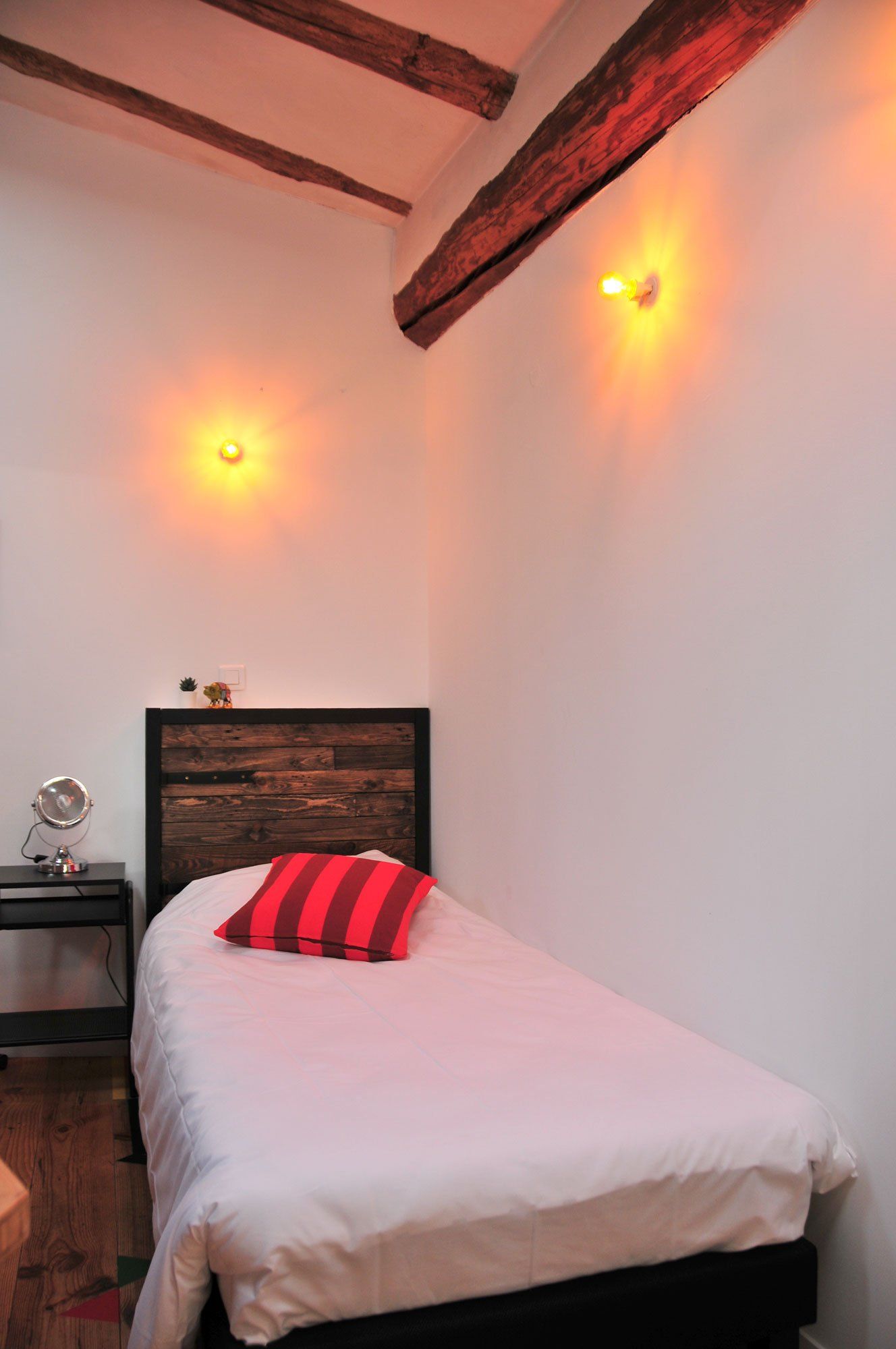 Chambre étroite et confortable avec un lit simple, un oreiller rouge, des appliques murales et des poutres apparentes au plafond.
