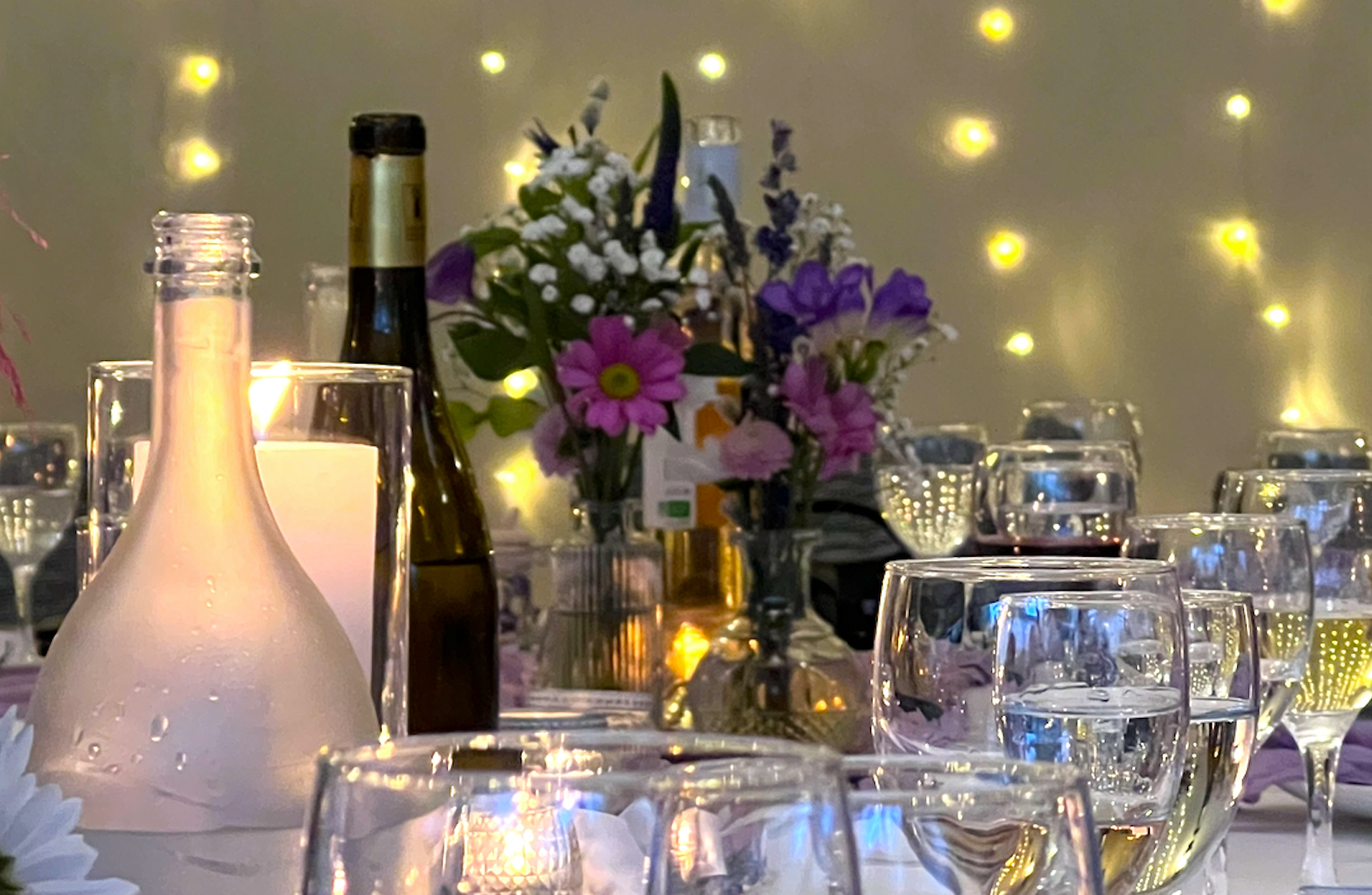 Une table élégamment dressée, illuminée de guirlandes lumineuses, ornée de fleurs, de verres et de bouteilles de vin.