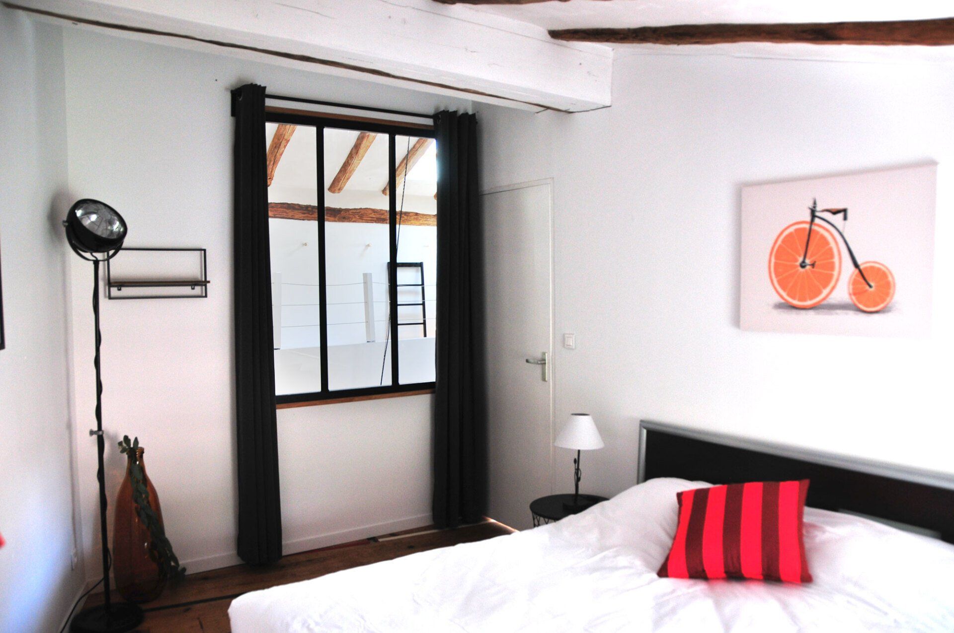 Chambre lumineuse avec fenêtre à cadre noir, lit blanc, oreiller à rayures rouges et tableau mural représentant un vélo orange