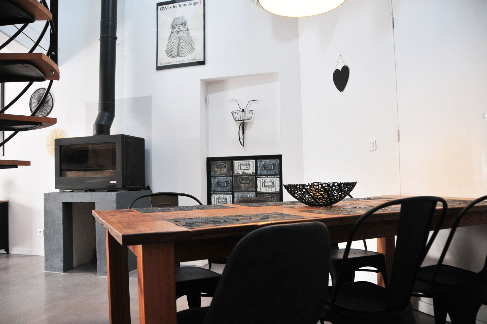 Salle à manger avec table en bois, chaises noires, poêle à bois et décorations murales dans un intérieur blanc lumineux