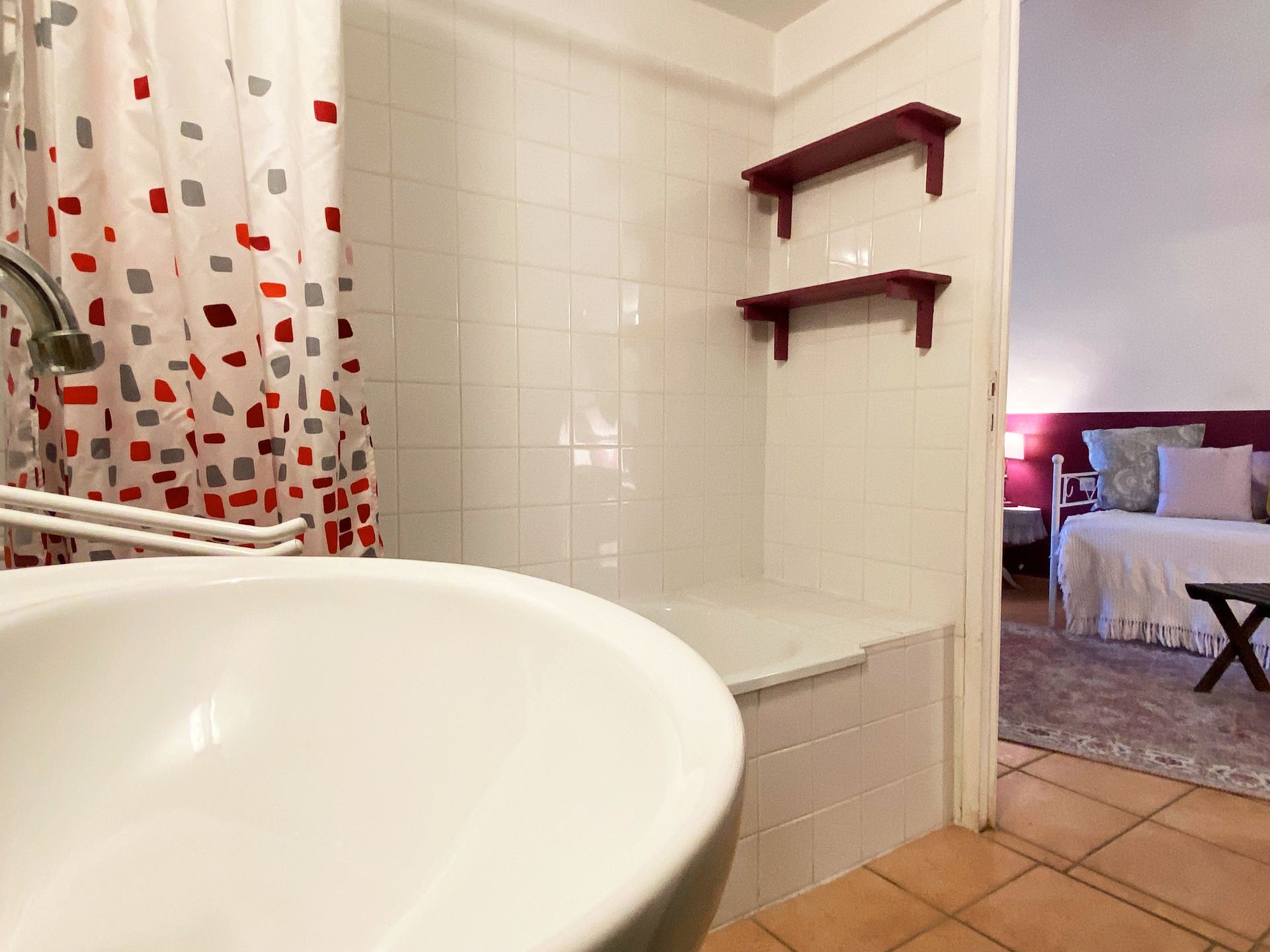 Petite salle de bains avec lavabo blanc, rideau de douche à motifs rouges, étagères murales et sol carrelé donnant sur une chambre.