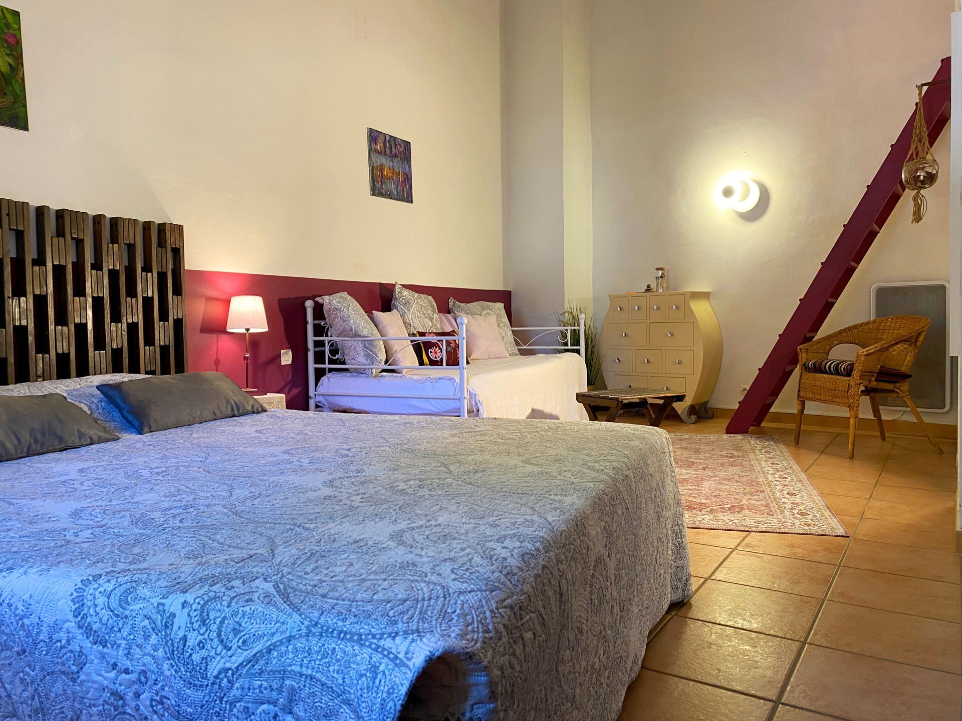 Chambre confortable avec un grand lit, une tête de lit éclairée en rose, un sol carrelé, une commode, une chaise et un plafond mansardé.