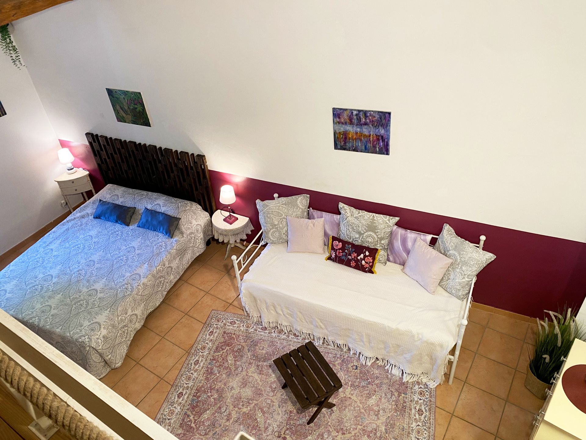 Petite chambre avec un lit et un canapé face à un tapis à motifs et une table basse, dans un décor blanc et bordeaux.