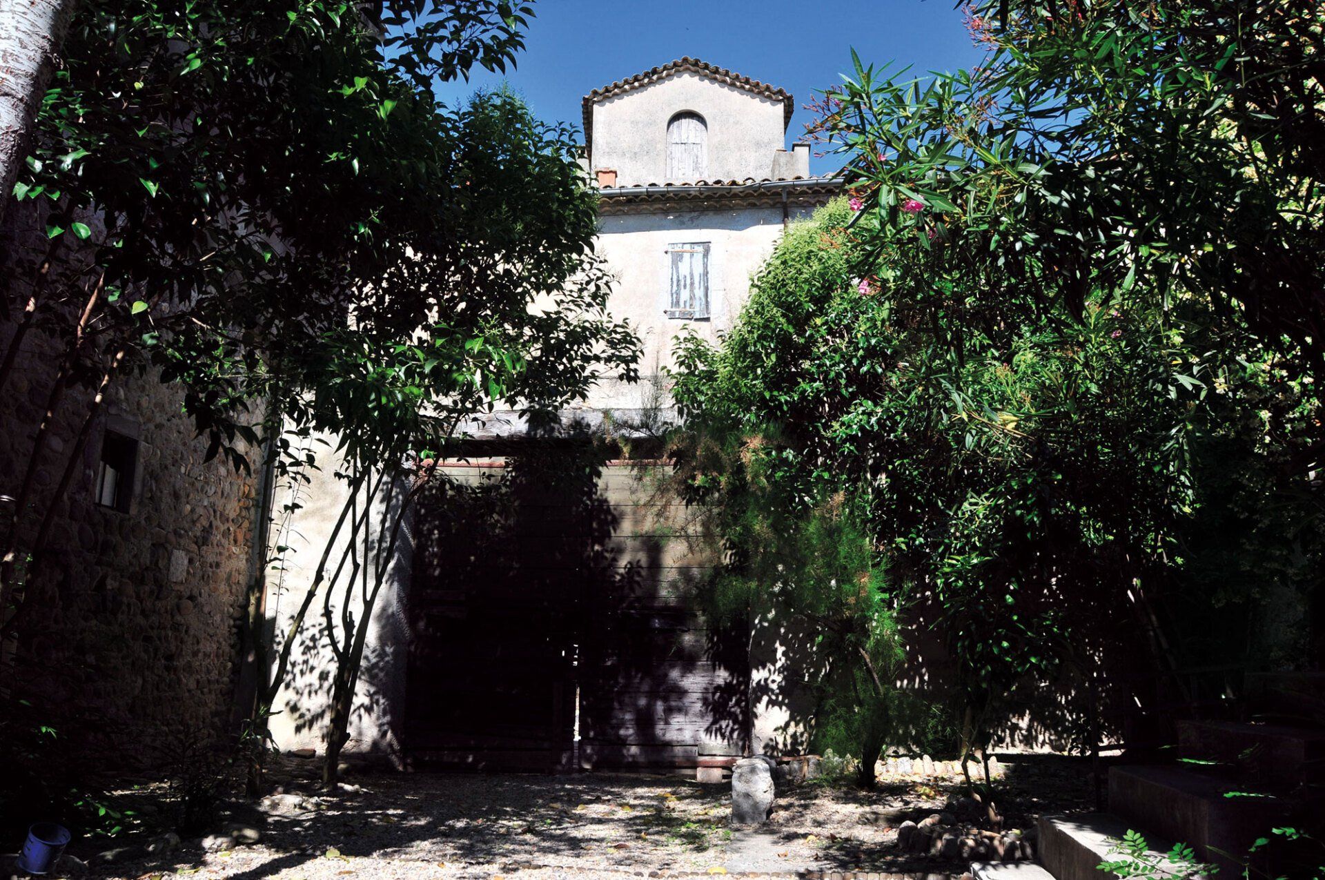 Bâtiment en pierre baigné de soleil, partiellement dissimulé par les arbres, avec une entrée voûtée sombre et une cour intérieure verdoyante.