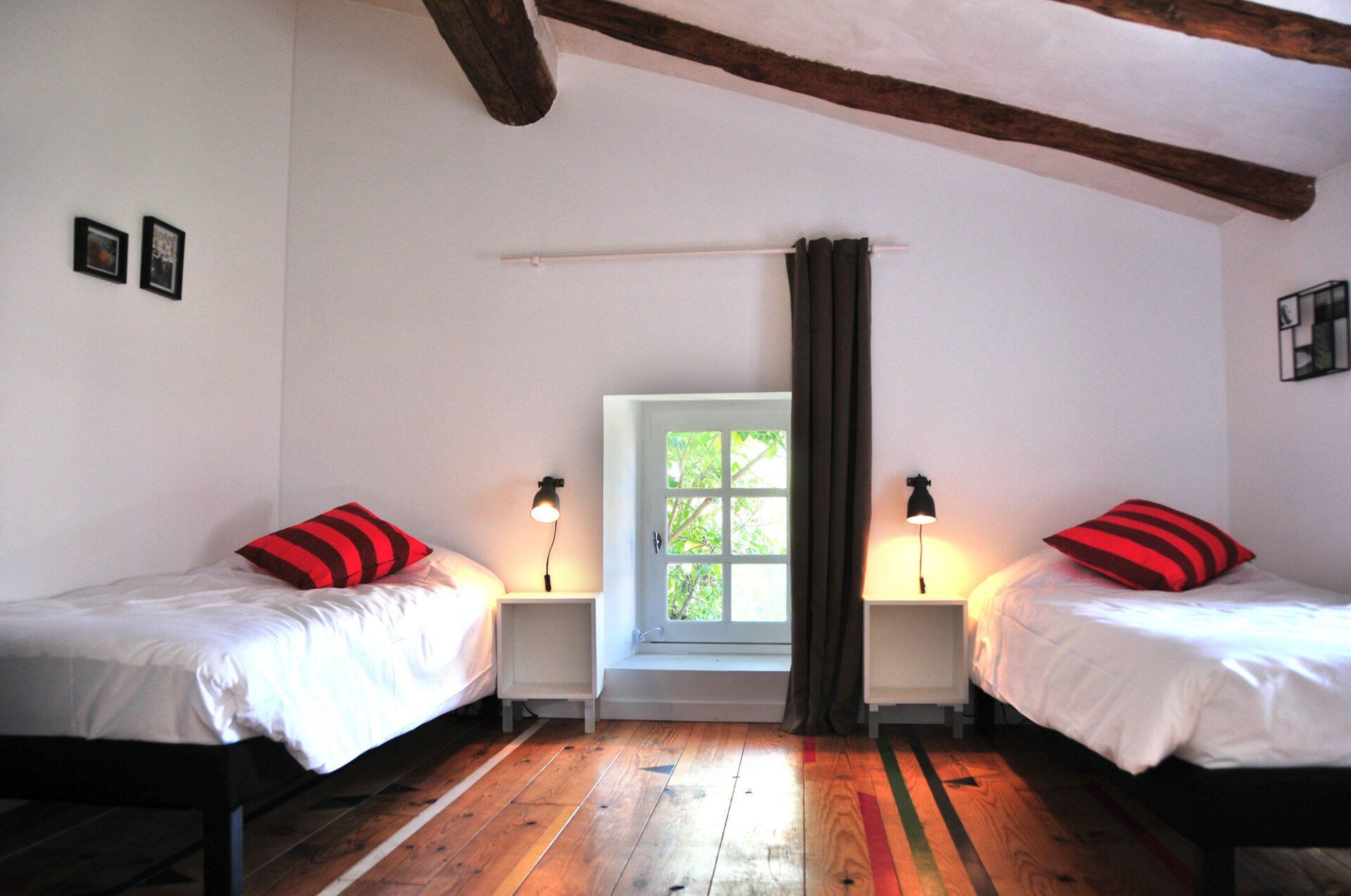 Chambre lumineuse avec deux lits jumeaux, murs blancs, parquet et une fenêtre entre les lampes et les rideaux.