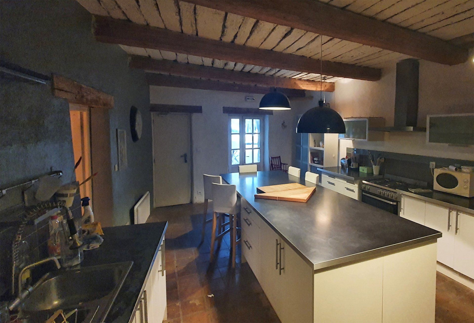 Cuisine rustique avec poutres apparentes, îlot central, luminaires suspendus et porte donnant sur une pièce lumineuse