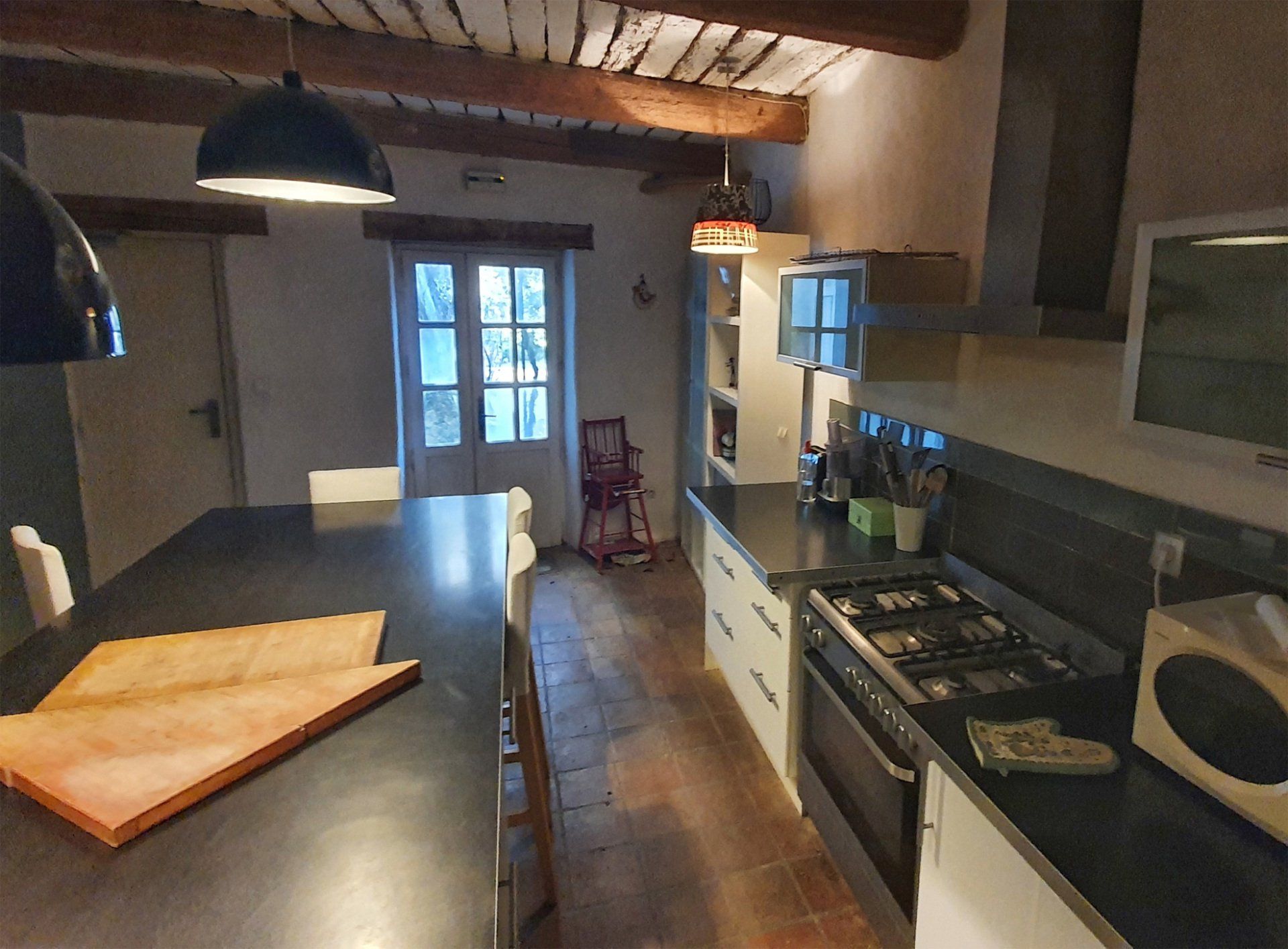 Cuisine rustique avec comptoirs noirs, poutres apparentes au plafond et porte donnant sur une terrasse arrière.