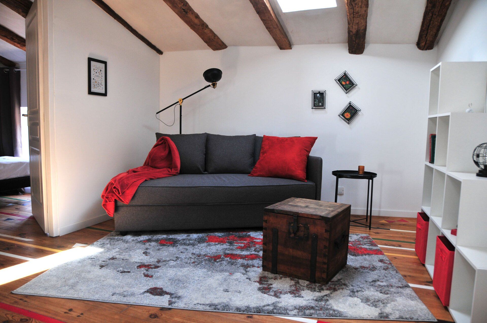 Salon cosy avec canapé gris, jeté rouge, tapis à motifs, poutres en bois et table basse cubique.