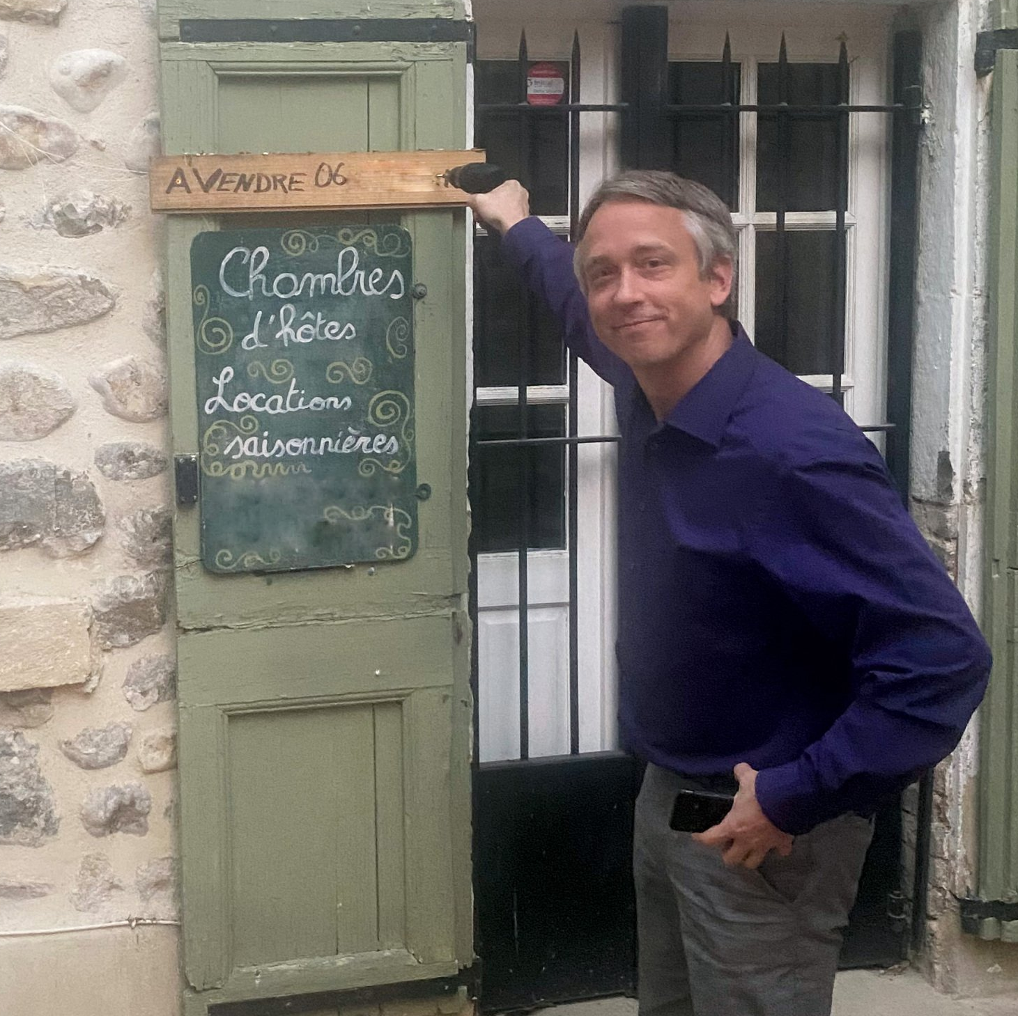 Mathijs désigne le menu d'un café sur un tableau noir, à côté d'une porte verte et d'un mur en pierre.