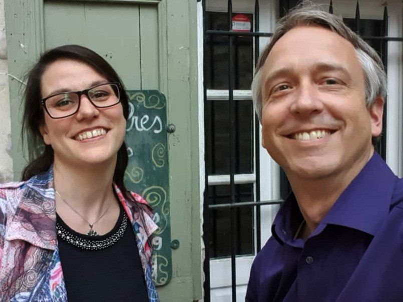 Deux personnes souriantes se tiennent devant un magasin avec une porte et une fenêtre vertes derrière elles.
