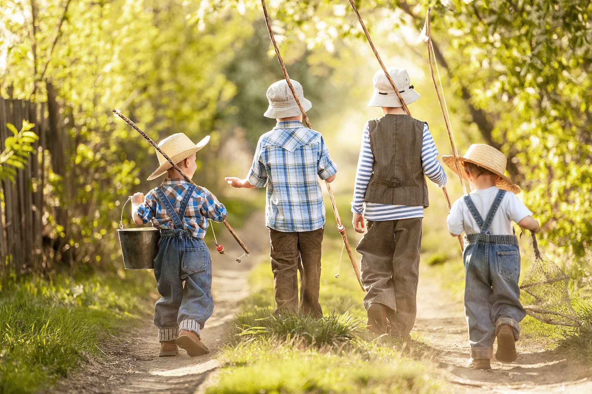 Quatre enfants coiffés de chapeaux de paille descendent un chemin de jardin ensoleillé, munis de cannes à pêche et de seaux.