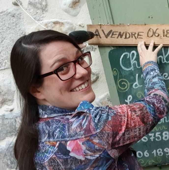 Sabine souriante portant des lunettes écrit sur un tableau noir vert à côté d'un mur en pierre