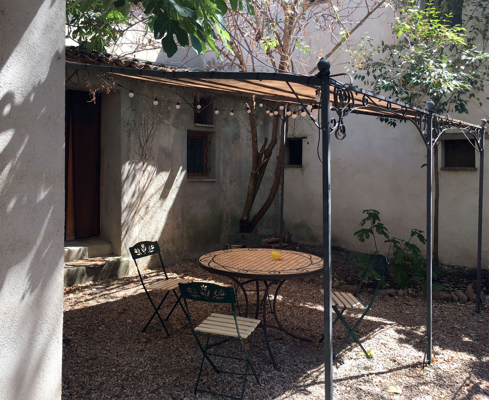 Patio ombragé avec une table ronde, des chaises et une pergola en métal à côté d'une maison en stuc