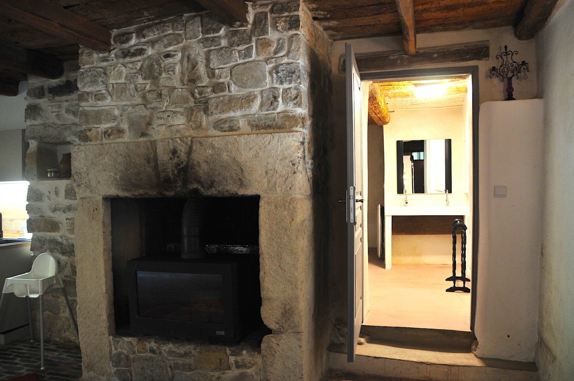 Cheminée en pierre et porte d'entrée dans une pièce rustique aux poutres apparentes, donnant sur un espace intérieur éclairé.