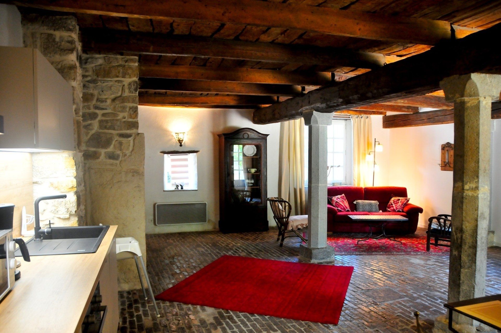 Salon rustique avec piliers en pierre, poutres en bois, tapis rouge et canapé rouge près de fenêtres lumineuses