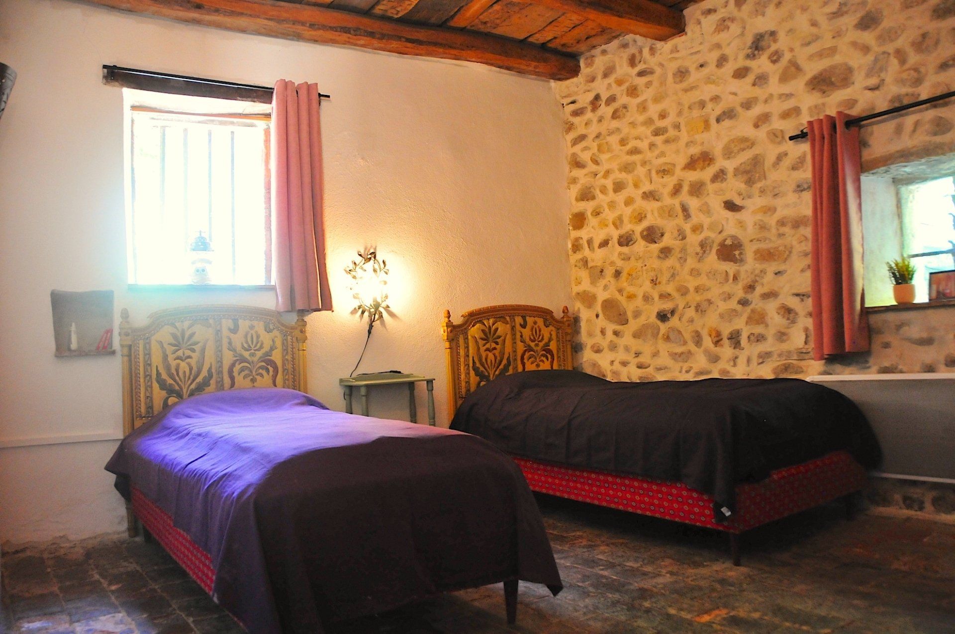 Chambre rustique avec deux lits, murs en pierre, rideaux roses et une fenêtre laissant entrer la lumière.