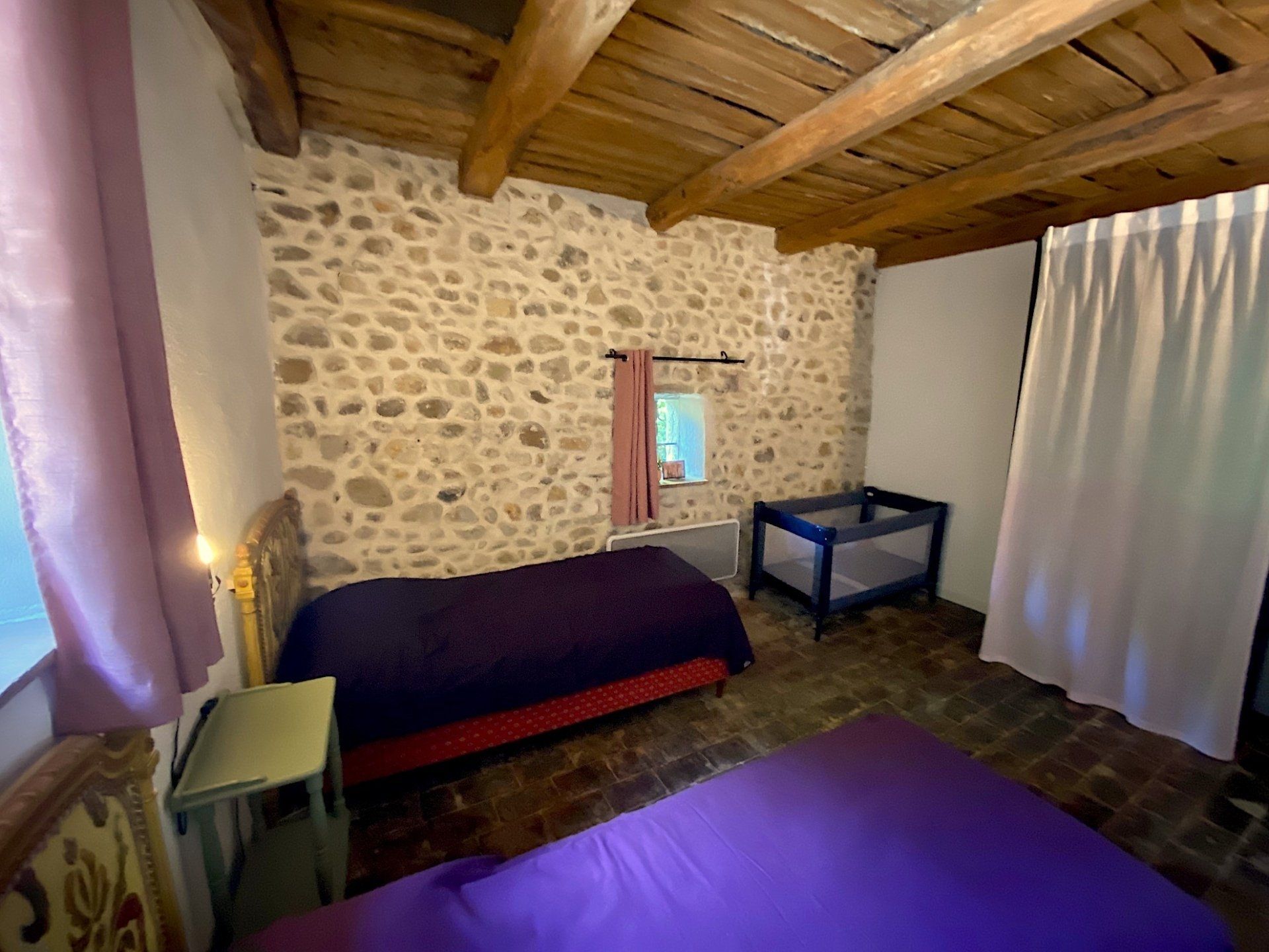 Chambre rustique avec murs en pierre, plafond en bois, lit, petite table et tapis violet