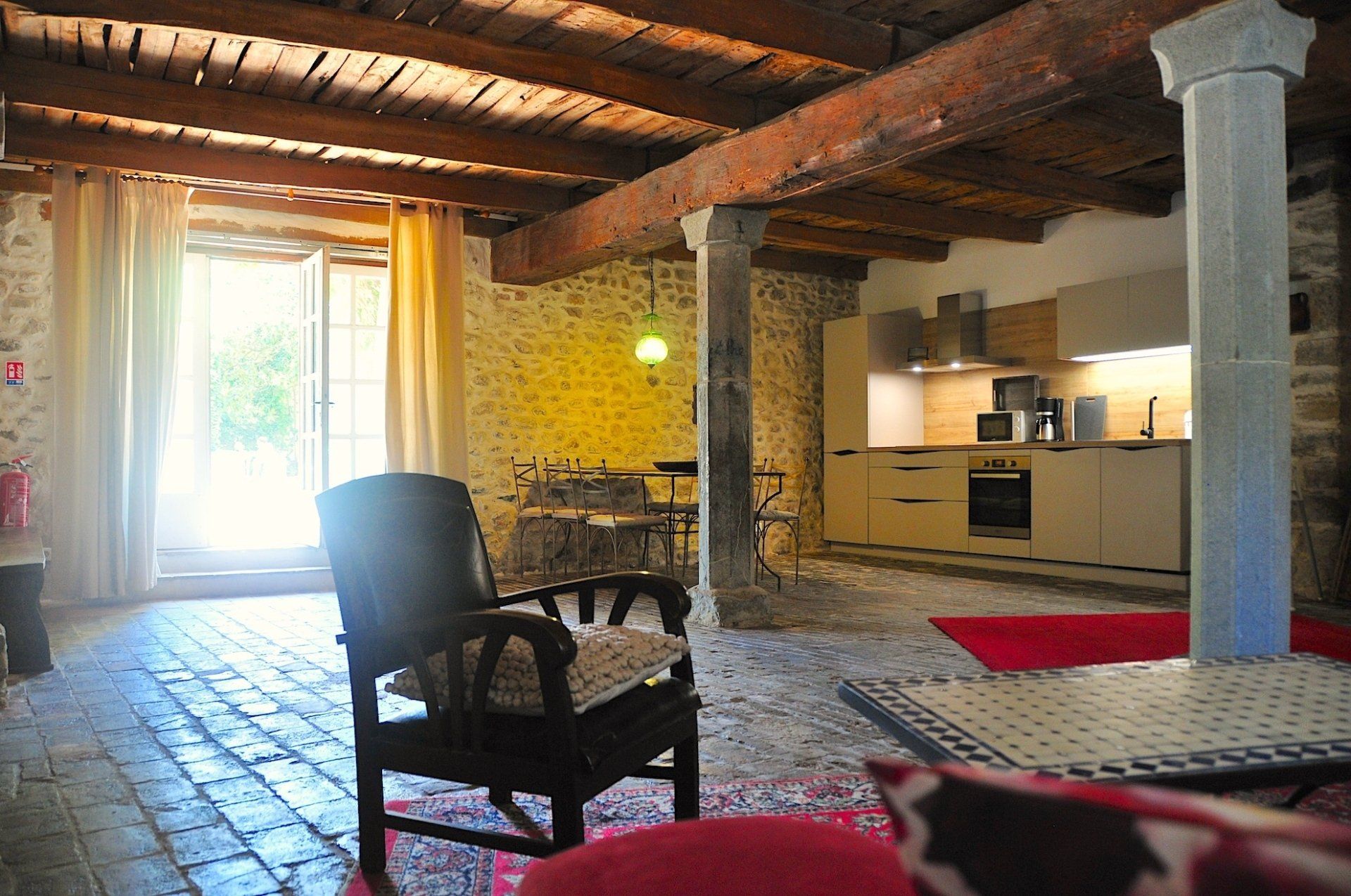 Salon rustique avec sol en pierre, poutres apparentes, fauteuil et fenêtre lumineuse jouxtant un petit coin cuisine.
