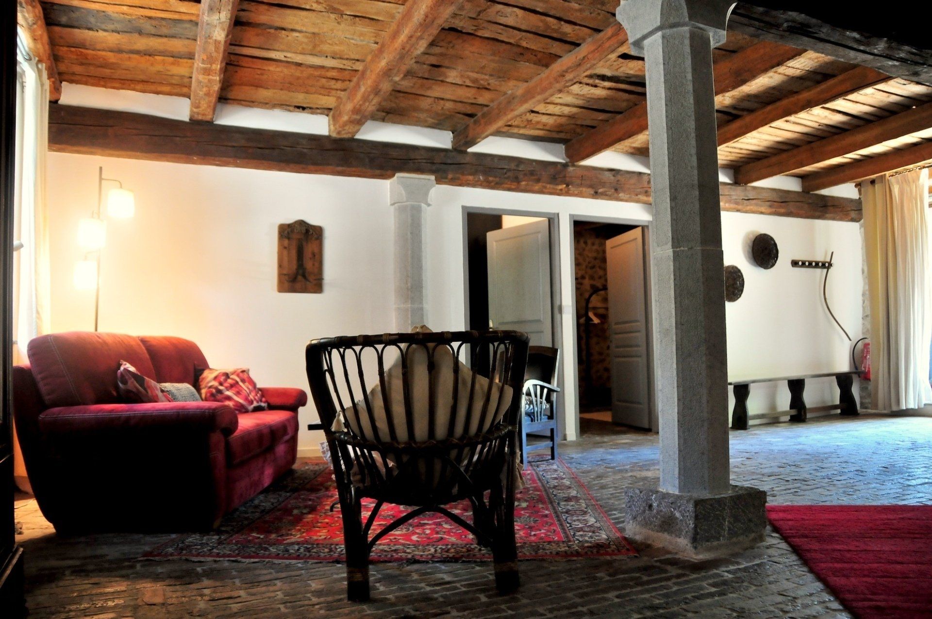 Salon rustique et chaleureux avec canapé rouge, fauteuil en osier, tapis à motifs et poutres apparentes.