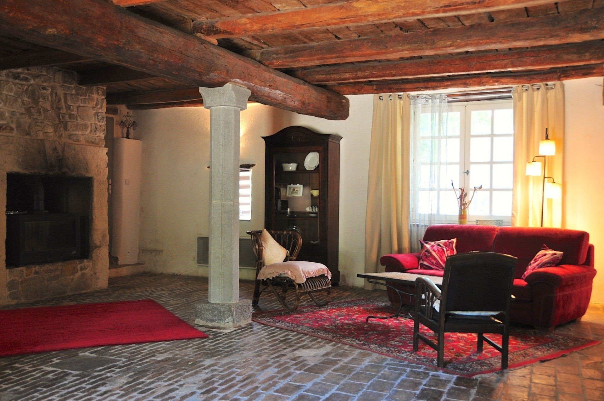 Salon rustique et chaleureux avec poutres apparentes, canapé rouge, cheminée et fenêtre baignée de soleil