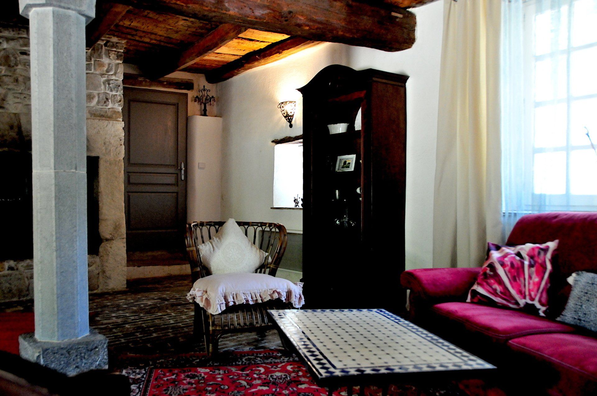Salon cosy avec canapé rouge, tapis à motifs, armoire haute et plafond aux poutres apparentes.