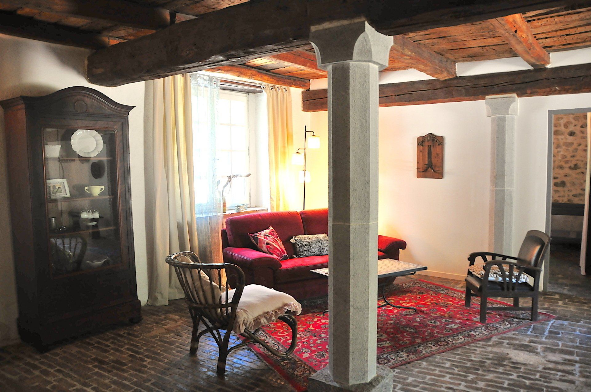 Salon rustique avec canapé rouge, tapis à motifs, poutres apparentes et vitrine près de la fenêtre