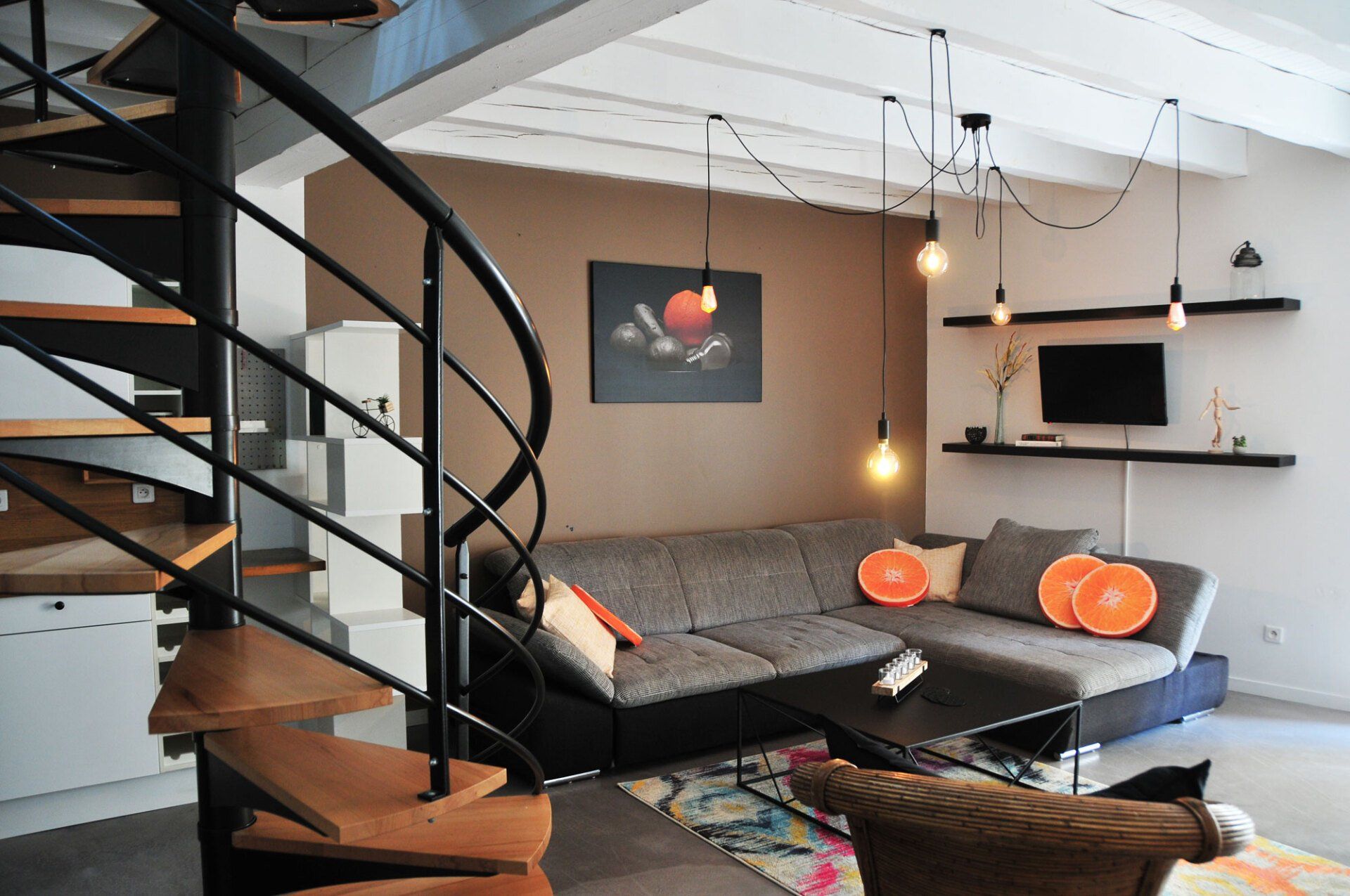 Salon loft moderne avec escalier en colimaçon, canapé d'angle noir, étagères murales et luminaires suspendus aux tons chauds.