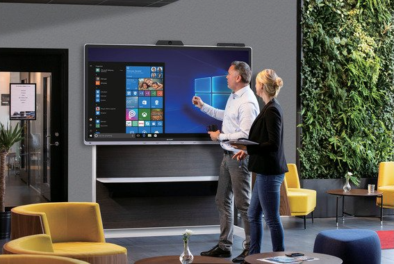 Dwie osoby stoją przed dużym ekranem wyświetlacza i wskazują na interfejs systemu Windows w biurze.